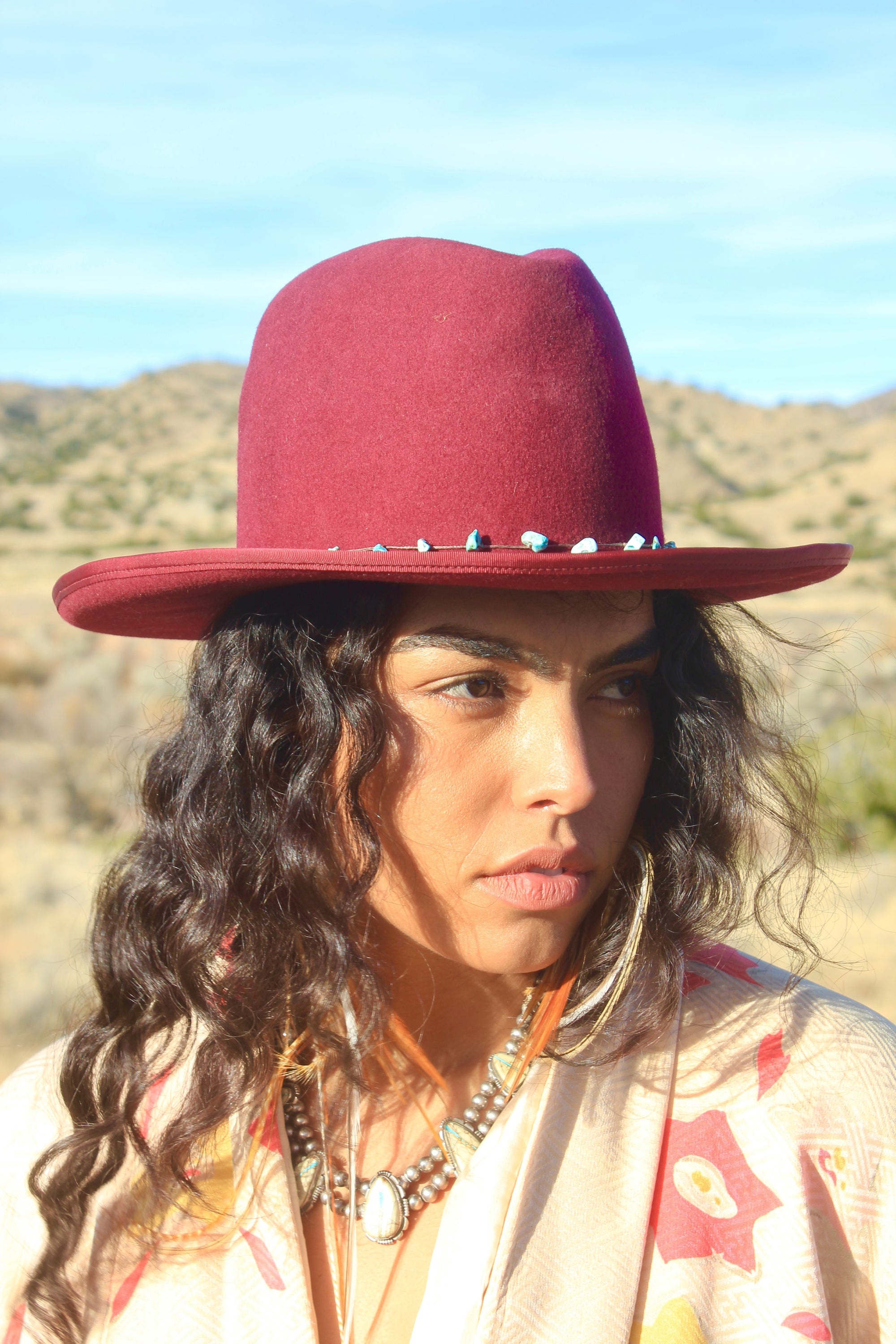 Left view of a woman wearing the LH Bordeaux Crease hat by Lone Hawk Hats featuring bordeaux fur blend felt, a slight crease crown, and a hand-tied turquoise band, size 7 1/4.LH Bordeaux Crease hat by Lone Hawk Hats featuring bordeaux fur blend felt, a slight crease crown, and a hand-tied turquoise band, size 7 1/4.