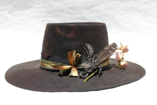 left view of The Emerald Hawk by Lone Hawk Hats, a distressed telescope crown western hat featuring a wide flat brim with Bardot dip, hand-dyed silk ribbon, dried flowers, and found feathers.